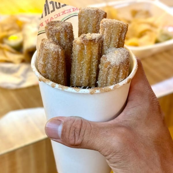A hand holds a white cup filled with several sugar-dusted churros from Adalbertacos Mexican Food.