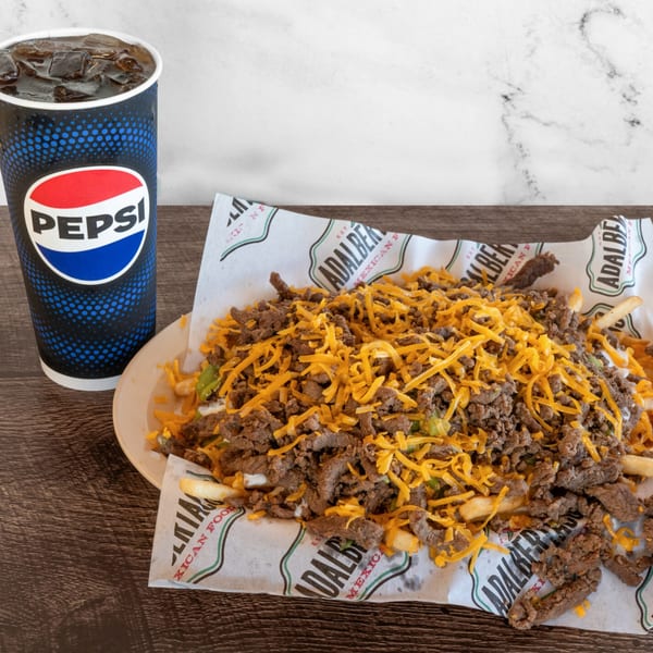 Adalbertacos carne asada fries, topped with shredded cheese and a Pepsi drink on a wooden table.